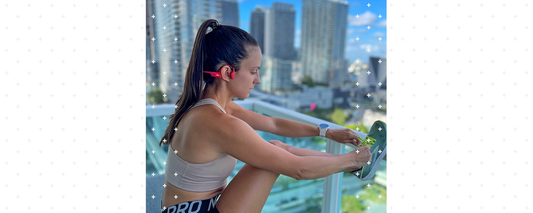 Woman standing on balcony doing leg stretches while wearing Shokz OpenRun wireless headphones